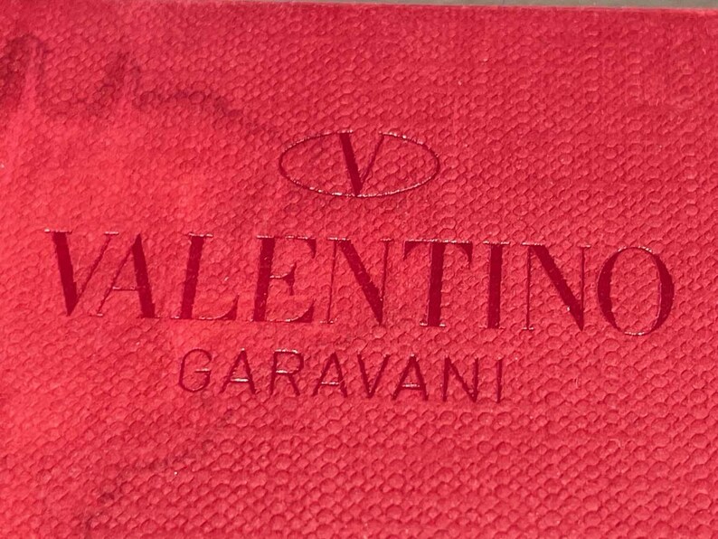 Puede incluir: Etiqueta de cuero roja con el texto "VALENTINO GARAVANI" en letras blancas y un logotipo "V" blanco en forma ovalada.