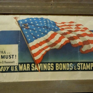 May include: A vintage World War II era poster featuring a waving American flag and the text "WE CAN... WE WILL... WE MUST! ... Franklin D. Roosevelt BUY U.S. WAR SAVINGS BONDS & STAMPS NOW".