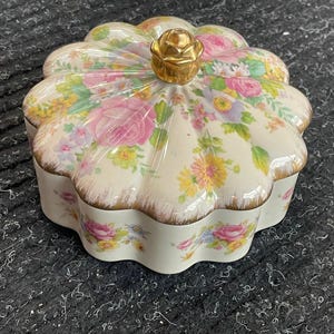 May include: A ceramic trinket box with a floral pattern featuring pink roses, yellow and purple flowers, and green leaves. The lid is topped with a gold-colored rose-shaped knob. The box has a scalloped edge with gold trim.