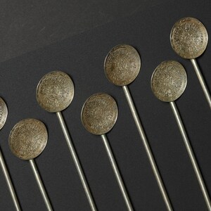 May include: Seven silver spoons with intricate engraved designs on the bowl and a long, thin handle. The spoons are arranged in a row on a black background.