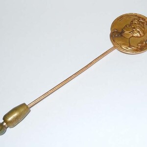 May include: A gold-toned pin with a round head featuring a raised design of a woman's face. The pin has a long, thin, gold-toned stem.