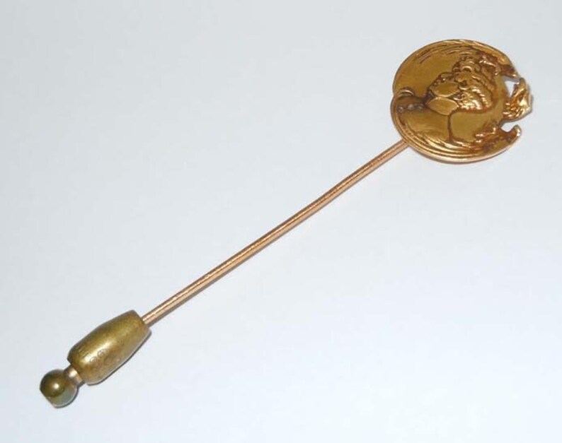 May include: A gold-toned pin with a round head featuring a raised design of a woman's face. The pin has a long, thin, gold-toned stem.