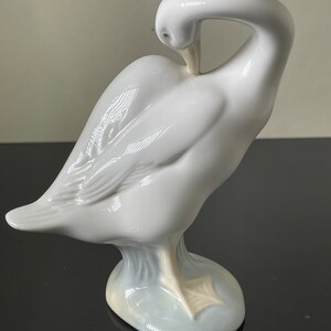 May include: A white porcelain figurine of a goose with its head tucked under its wing. The goose is standing on a light blue base.