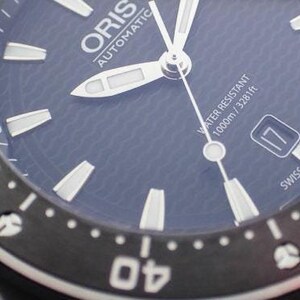 Puede incluir: Primer plano de una esfera de reloj autom&aacute;tico Oris negra con marcas blancas. El reloj es resistente al agua hasta 1000 m / 3281 pies. La esfera del reloj tiene una esfera azul con un patr&oacute;n de ondas.