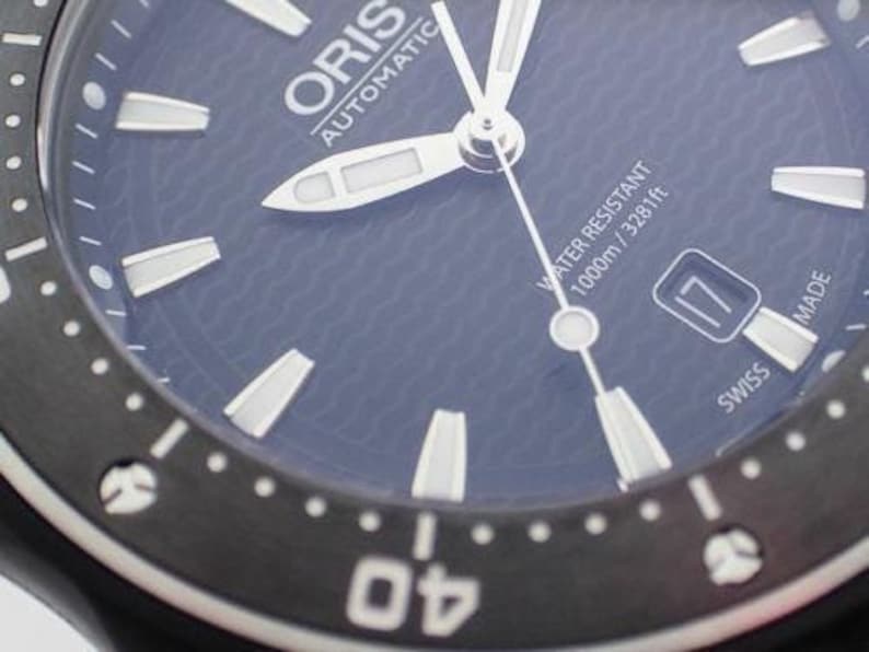 Puede incluir: Primer plano de una esfera de reloj autom&aacute;tico Oris negra con marcas blancas. El reloj es resistente al agua hasta 1000 m / 3281 pies. La esfera del reloj tiene una esfera azul con un patr&oacute;n de ondas.