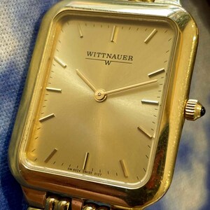 May include: A gold-toned Wittnauer wristwatch with a rectangular face and a matching gold band. The watch face displays the brand name and the letter "W". The watch has a gold-colored dial with gold hands and hour markers.
