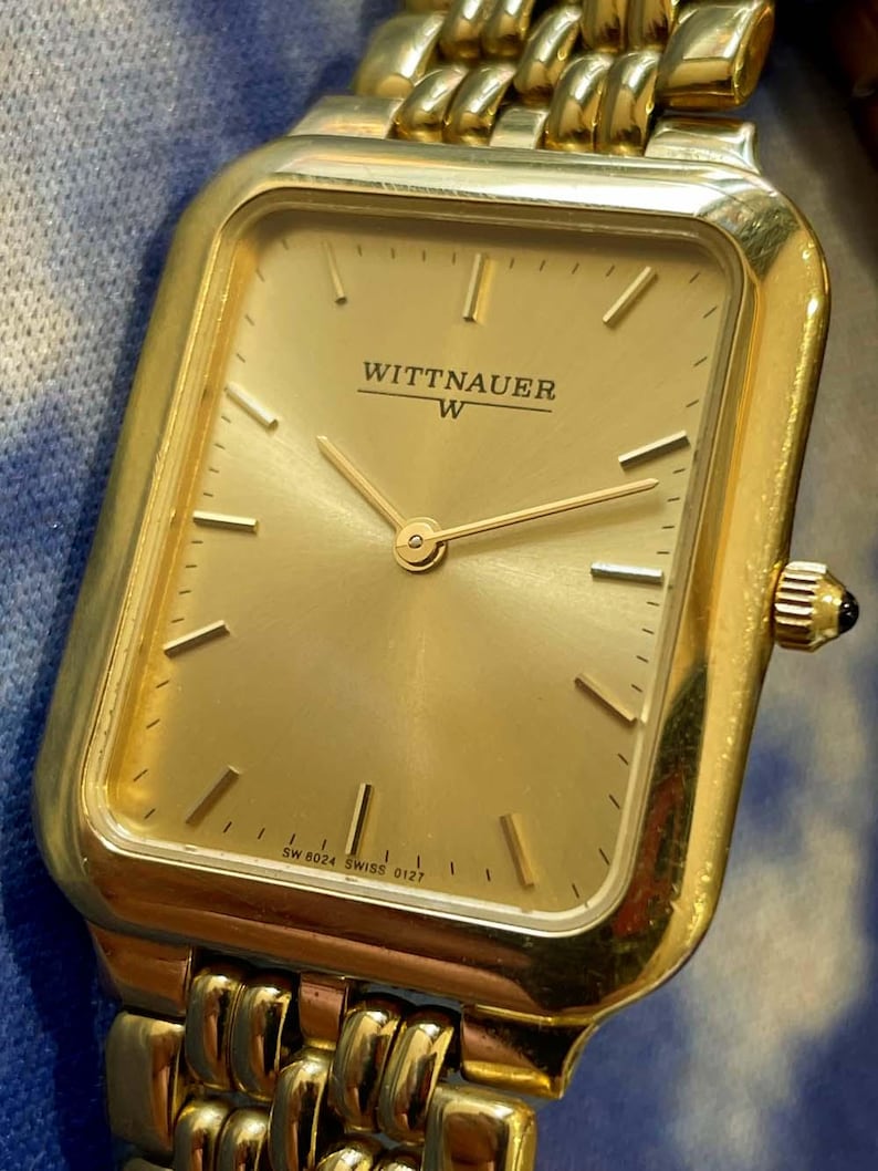 May include: A gold-toned Wittnauer wristwatch with a rectangular face and a matching gold band. The watch face displays the brand name and the letter "W". The watch has a gold-colored dial with gold hands and hour markers.