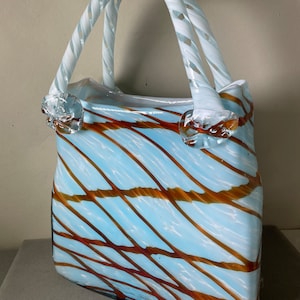May include: A white glass handbag with a blue and brown striped pattern. The handles are twisted and white.