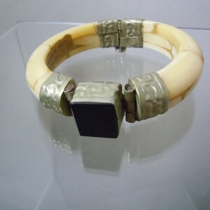 May include: A white bangle bracelet with silver accents and a black square clasp. The bracelet is made of bone and has a carved design.