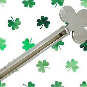 Puede incluir: Una llave plateada con forma de trébol, probablemente para un mecanismo de cuerda, sobre un fondo blanco con tréboles verdes dispersos. La llave tiene un cuerpo largo y cilíndrico con una ranura. La cabeza en forma de trébol está en la parte superior.