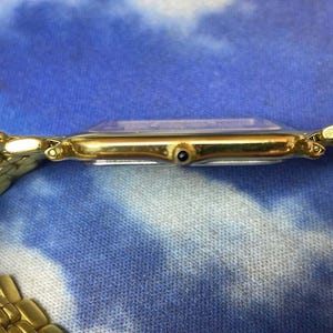 May include: A gold-toned wristwatch with a rectangular face and a matching gold link band. The watch face is clear, and the band is made of small, connected links. The watch is set against a blue and white patterned background.