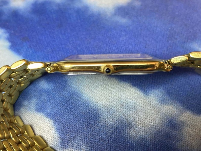 May include: A gold-toned wristwatch with a rectangular face and a matching gold link band. The watch face is clear, and the band is made of small, connected links. The watch is set against a blue and white patterned background.