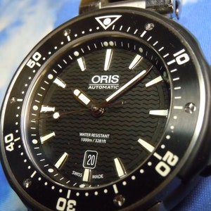 Puede incluir: Un reloj autom&aacute;tico Oris negro con una esfera blanca y agujas plateadas. El reloj es resistente al agua hasta 1000 metros o 3281 pies. El reloj tiene una ventana de fecha en la posici&oacute;n de las 3 en punto y un bisel negro con marcas blancas.