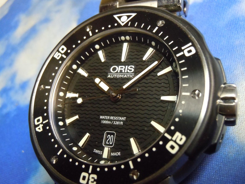 Puede incluir: Un reloj autom&aacute;tico Oris negro con una esfera blanca y agujas plateadas. El reloj es resistente al agua hasta 1000 metros o 3281 pies. El reloj tiene una ventana de fecha en la posici&oacute;n de las 3 en punto y un bisel negro con marcas blancas.