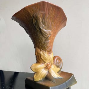 May include: A brown ceramic vase with a flared top and a square base. The vase features a floral design with a large yellow flower and a snail. The vase is approximately 10 inches tall.