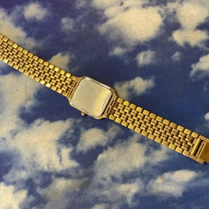 May include: A gold-toned wristwatch with a rectangular face and a linked band. The watch is displayed against a blue background with a cloud pattern. The watch band is approximately 7 inches long.