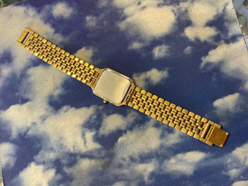 May include: A gold-toned wristwatch with a rectangular face and a linked band. The watch is displayed against a blue background with a cloud pattern. The watch band is approximately 7 inches long.
