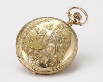 May include: A gold pocket watch with intricate floral and bird engravings. The watch features a detailed design with a bird in flight and delicate flowers in shades of gold, white, and pink. The watch has a winding stem and a circular bail.