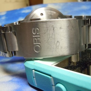 Puede incluir: Un reloj de pulsera plateado con una correa de acero inoxidable. La esfera del reloj no es visible. La correa tiene la palabra "ORIS" grabada.