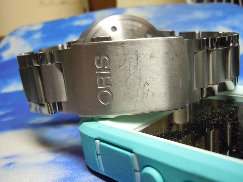 Puede incluir: Un reloj de pulsera plateado con una correa de acero inoxidable. La esfera del reloj no es visible. La correa tiene la palabra "ORIS" grabada.