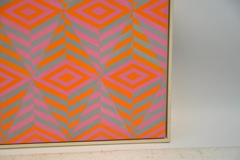 May include: Abstract geometric artwork with a repeating pattern of diamonds and chevrons. The design uses orange, pink, and grey, creating a vibrant visual effect. The artwork is framed.