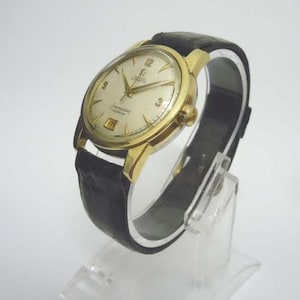 Könnte beinhalten: Eine Vintage Omega Seamaster Calendar Uhr in Goldoptik mit cremefarbenem Zifferblatt und schwarzem Lederarmband. Die Uhr hat goldene Zeiger und Stundenmarkierungen sowie ein Datumsfenster. Die Uhr wird auf einem transparenten Ständer präsentiert.
