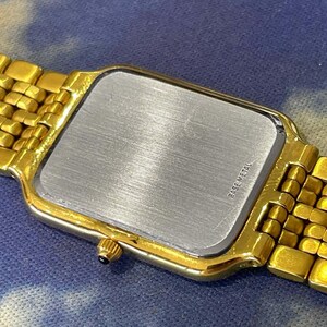 May include: Close-up of a gold-toned wristwatch with a rectangular face and a matching gold band. The watch's back is silver with the words "BASE METAL" printed on it. The watch is on a blue and white background.