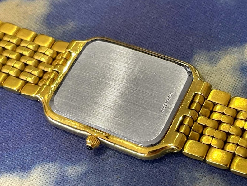 May include: Close-up of a gold-toned wristwatch with a rectangular face and a matching gold band. The watch's back is silver with the words "BASE METAL" printed on it. The watch is on a blue and white background.