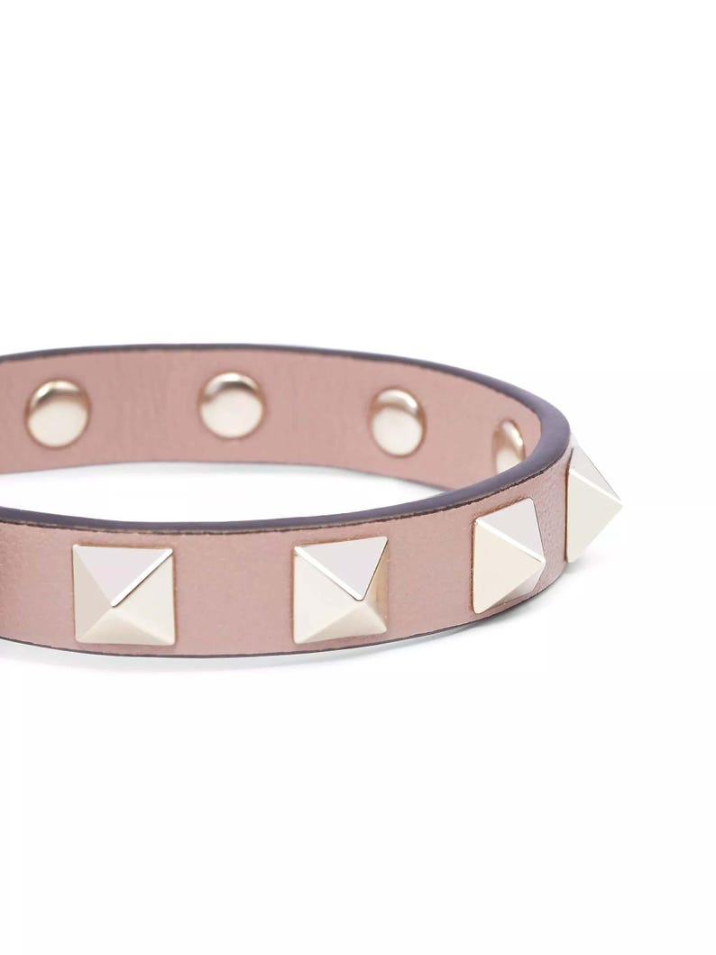 Puede incluir: Un brazalete de cuero rosa claro con tachuelas de pir&aacute;mide plateadas.