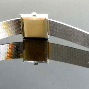 May include: A silver watch with a square face and a metal band. The watch is on a reflective surface.