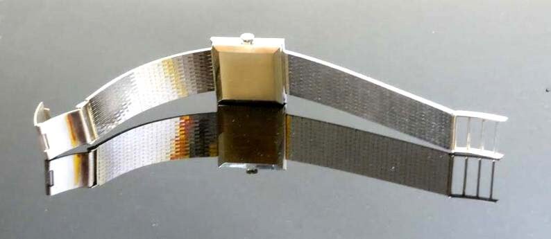 May include: A silver watch with a square face and a metal band. The watch is on a reflective surface.