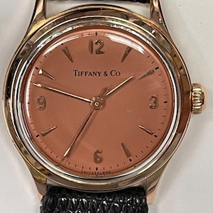May include: A gold-tone wristwatch with a black leather strap. The watch face is a light brown colour with black Roman numeral hour markers and black hands. The watch face also has the text "Tiffany & Co" and "SWITZERLAND" printed on it.