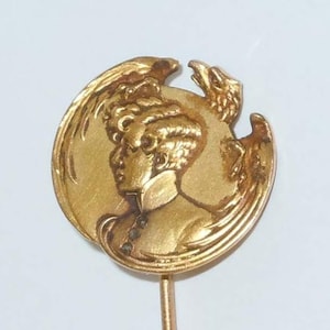 May include: A gold-toned pin with a round design featuring a profile of a woman with a bird behind her. The pin is attached to a thin gold-toned stem.