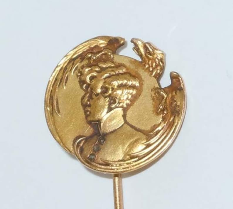 May include: A gold-toned pin with a round design featuring a profile of a woman with a bird behind her. The pin is attached to a thin gold-toned stem.