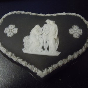 May include: Black and white ceramic heart-shaped trinket box with a raised design of three figures in classical dress. The box is decorated with a floral border.