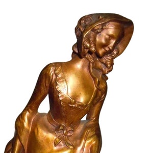 May include: A gold-toned metal sculpture of a woman in a dress and hat. The woman is looking down and her head is tilted to the side.