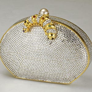 Op de afbeelding: Ovale, zilverkleurige clutch bedekt met kleine, sprankelende kristallen. De clutch heeft een goudkleurig frame en een decoratief, met juwelen bezet rupsontwerp aan de bovenkant, met een parelaccent. Een luxe avondtas.