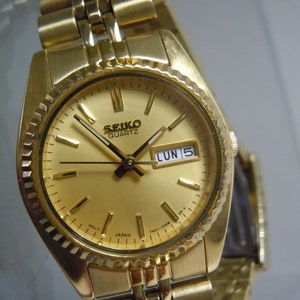 May include: A gold-toned Seiko Quartz watch with a matching gold band. The watch face displays the day and date, with the day reading "LUN 5". The watch has a textured bezel and a gold dial with gold hands and markers.