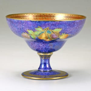 May include: A blue footed bowl with a speckled texture and gold trim. The bowl features a painted design of colorful fruit and leaves. The rim has a gold geometric pattern. The base and stem are also blue with gold accents.