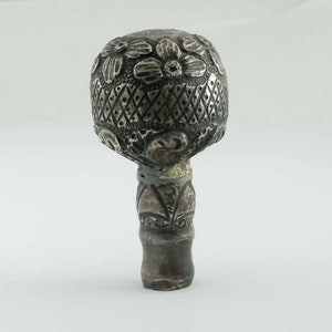 Op de afbeelding: Een zilveren flessenstop met een ingewikkeld bloemmotief. De bovenkant van de stop is rond en heeft een herhalend patroon van bloemen en diamanten. De onderkant van de stop is cilindrisch en heeft een vergelijkbaar bloemmotief.