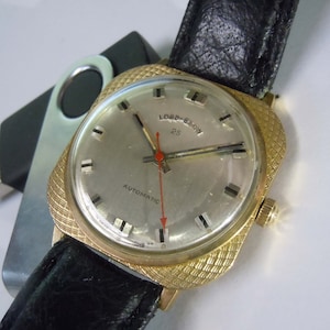 Op de afbeelding: Een gouden polshorloge met een vierkante wijzerplaat en een zwarte leren band. De wijzerplaat heeft een witte achtergrond met zwarte markeringen en de tekst "Lord Elgin 25 Automatic".