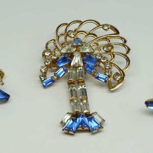 May include: A gold tone brooch and matching earrings set with blue and clear rhinestones. The brooch features a stylized tree design with a fan-shaped top.