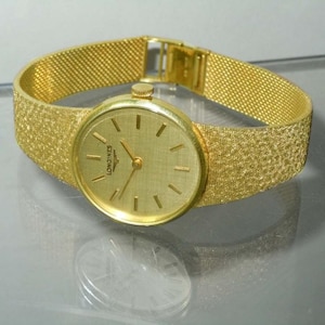 May include: A gold-tone Longines wristwatch with a gold mesh band. The watch face is round and has a white background with black Roman numeral hour markers and black hands.