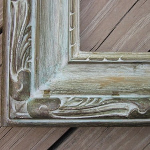 May include: A close-up of a distressed, light green picture frame with ornate detailing. The frame has a weathered, aged look with visible brushstrokes and a hint of gold.