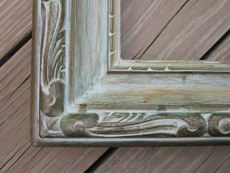 May include: A close-up of a distressed, light green picture frame with ornate detailing. The frame has a weathered, aged look with visible brushstrokes and a hint of gold.