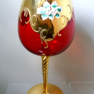 May include: A red glass wine goblet with a gold rim and a gold twisted stem. The goblet is decorated with a gold floral design and a white flower with green leaves.