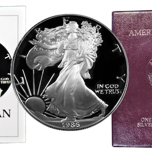 Könnte beinhalten: Eine silberne American Eagle Proof-Münze aus dem Jahr 1986 in einer roten Samtbox mit den Worten "American Eagle" und "One Ounce Proof Silver Bullion Coin" auf der Vorderseite. Die Münze zeigt eine Darstellung von Lady Liberty, die mit einer Fackel und einem Olivenzweig geht.