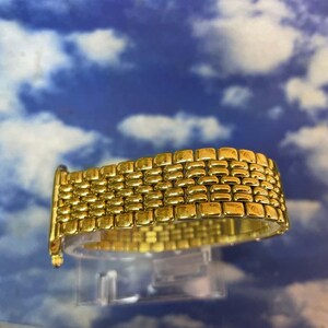 May include: A gold-toned watch band with a woven design. The band is made of small, rectangular links that are closely connected. The watch band is displayed against a blue and white cloudy background.