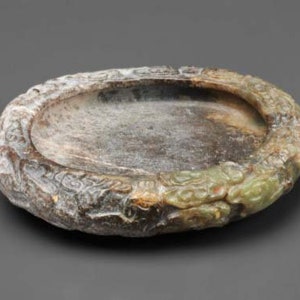 May include: A round, shallow dish carved from jade with a green and brown mottled color. The dish has a raised rim with intricate carvings of dragons and other mythical creatures.