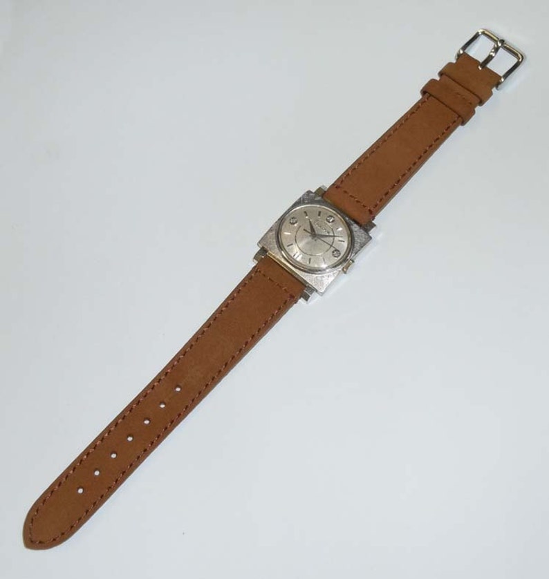 May include: A silver square-shaped wristwatch with a white face and black hands. The watch has a brown leather band with a buckle closure.
